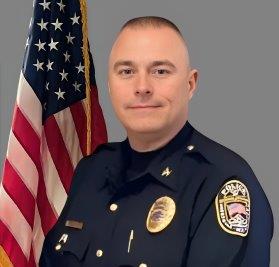Portrait of a man in a dark police uniform with a badge, shoulder patch, and gold pins. He has short-cropped hair and stands in front of the American flag, facing slightly right. Neutral gray background.