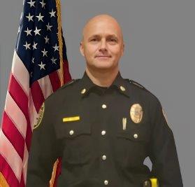 'Portrait of a bald man in a black police uniform with a badge, shoulder patch, and gold pins. He stands with a neutral expression in front of the American flag. Neutral gray background.'