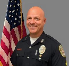 'Portrait of a bald man in a black police uniform with a badge and several pins. He is smiling and standing in front of the American flag. Neutral gray background.'