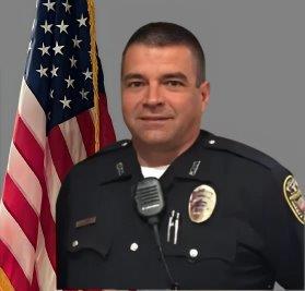 Portrait of a man in a black police uniform with a badge and a radio microphone clipped to his shirt. He stands in front of the American flag and is smiling slightly. Neutral gray background.