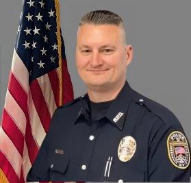 Portrait of a man in a dark police uniform with badge, shoulder patch, and two pens in his pocket. He has short, light-colored hair and is smiling. The American flag is visible behind him. Neutral gray background.