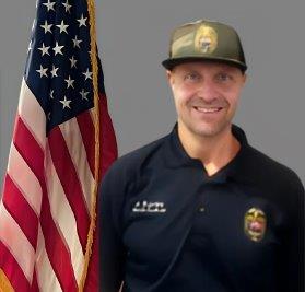 Portrait of a man with a light complexion, wearing a camo hat and a navy blue police polo shirt with a badge. He is smiling and stands in front of the American flag. Neutral gray background.