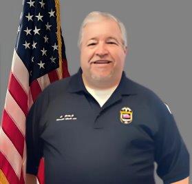 Portrait of a smiling man with short, white hair, wearing a navy blue police polo shirt with badge and name embroidery. He stands in front of the American flag. Neutral gray background.