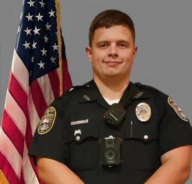 Portrait of a man with short brown hair in a black police uniform with badge, name tag, shoulder patch, and body camera. He is standing in front of the American flag, arms folded. Gray background.
