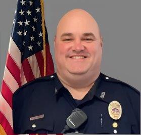 Portrait of a bald man in a dark police uniform with a badge and radio microphone. He is smiling, standing in front of the American flag with a gray background.