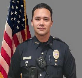 Portrait of a man with dark hair in a black police uniform with badge, name tag, shoulder patch, and police radio. He is standing in front of the American flag with a neutral expression. Gray background.