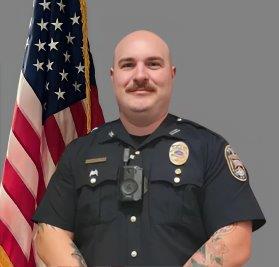 Portrait of a man with a shaved head and mustache in a black police uniform with badge, shoulder patch, and body camera. He is standing in front of the American flag. Gray background.