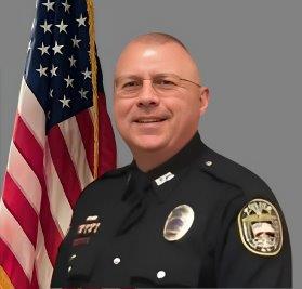 Portrait of a man with short, light hair and glasses in a black police uniform with badge and shoulder patch. He is smiling and standing in front of the American flag with a gray background.