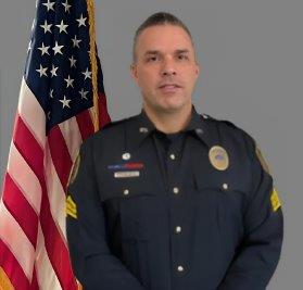 Portrait of a man with short dark hair in a black police uniform with badge, shoulder patch, and sergeant stripes on his sleeves. He stands in front of the American flag with a neutral expression. Gray background.