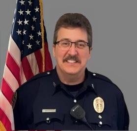 Portrait of a man with short dark hair, mustache, and glasses in a black police uniform with badge and radio microphone. He is smiling and standing in front of the American flag. Gray background.