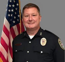 Portrait of a man with short light hair in a black police uniform with badge and name tag. He is smiling and standing in front of the American flag. Gray background.