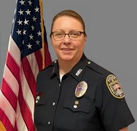 Portrait of a woman with light brown hair in a black police uniform with badge, shoulder patch, and glasses. She is smiling and standing in front of the American flag. Gray background.