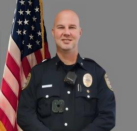 Portrait of a bald man in a black police uniform with badge, shoulder patch, and body camera. He is smiling and standing in front of the American flag. Gray background.