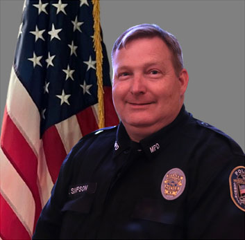 Portrait of a man with short hair in a dark police uniform with a badge, name tag, and sergeant stripes on his sleeve. He is standing in front of the American flag and has a neutral expression. Gray background.