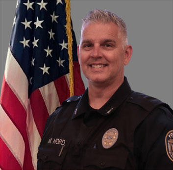 Portrait of a man with short gray hair in a black police uniform with a badge, pen, and name tag. He is smiling and standing in front of the American flag. Gray background.