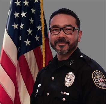 Portrait of a bald man in a dark police uniform with a badge and radio microphone. He is smiling, standing in front of the American flag with a gray background.