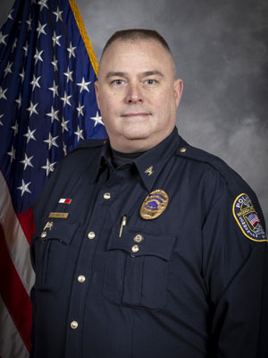 Portrait of a man in a dark police uniform with a badge, shoulder patch, and gold pins. He has short-cropped hair and stands in front of the American flag, facing slightly right. Neutral gray background.