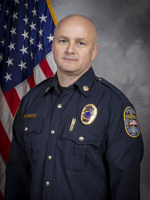 'Portrait of a bald man in a black police uniform with a badge, shoulder patch, and gold pins. He stands with a neutral expression in front of the American flag. Neutral gray background.'