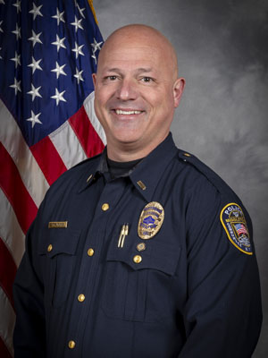 'Portrait of a bald man in a black police uniform with a badge and several pins. He is smiling and standing in front of the American flag. Neutral gray background.'