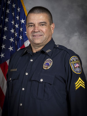 Portrait of a man in a black police uniform with a badge and a radio microphone clipped to his shirt. He stands in front of the American flag and is smiling slightly. Neutral gray background.