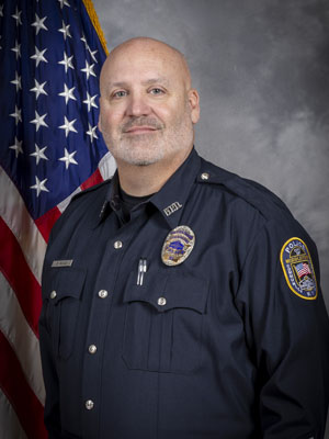 Portrait of a man in a black police uniform with a badge and a radio microphone clipped to his shirt. He stands in front of the American flag and is smiling slightly. Neutral gray background.