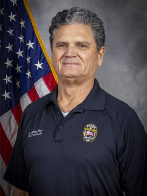 Portrait of a man with a light complexion, wearing a camo hat and a navy blue police polo shirt with a badge. He is smiling and stands in front of the American flag. Neutral gray background.