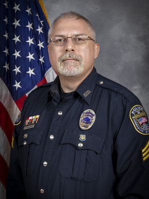 Portrait of a man with short, light hair and glasses in a black police uniform with badge and shoulder patch. He is smiling and standing in front of the American flag with a gray background.