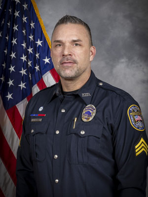 Portrait of a man with short dark hair in a black police uniform with badge, shoulder patch, and sergeant stripes on his sleeves. He stands in front of the American flag with a neutral expression. Gray background.