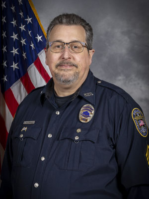 Portrait of a man with short dark hair, mustache, and glasses in a black police uniform with badge and radio microphone. He is smiling and standing in front of the American flag. Gray background.