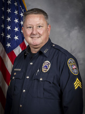 Portrait of a man with short light hair in a black police uniform with badge and name tag. He is smiling and standing in front of the American flag. Gray background.