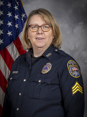 Portrait of a woman with light brown hair in a black police uniform with badge, shoulder patch, and glasses. She is smiling and standing in front of the American flag. Gray background.