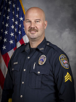 Portrait of a bald man in a black police uniform with badge, shoulder patch, and body camera. He is smiling and standing in front of the American flag. Gray background.