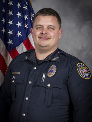 Portrait of a man with short brown hair in a black police uniform with badge, name tag, shoulder patch, and body camera. He is standing in front of the American flag, arms folded. Gray background.
