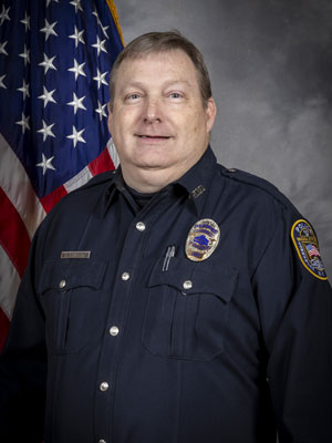 Portrait of a man with short hair in a dark police uniform with a badge, name tag, and sergeant stripes on his sleeve. He is standing in front of the American flag and has a neutral expression. Gray background.