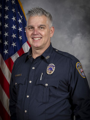 Portrait of a man with short gray hair in a black police uniform with a badge, pen, and name tag. He is smiling and standing in front of the American flag. Gray background.