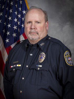 Portrait of a man with light brown hair in a black police uniform with badge and shoulder patch, standing with hands folded in front of the American flag. He has a serious expression. Gray background.