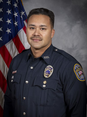 Portrait of a man with dark hair in a black police uniform with badge, name tag, shoulder patch, and police radio. He is standing in front of the American flag with a neutral expression. Gray background.