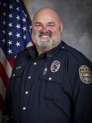 Portrait of a bald man in a dark police uniform with a badge and radio microphone. He is smiling, standing in front of the American flag with a gray background.