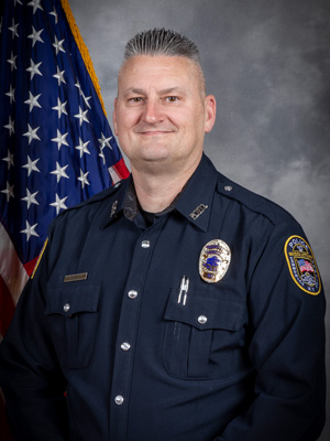 Portrait of a man in a dark police uniform with badge, shoulder patch, and two pens in his pocket. He has short, light-colored hair and is smiling. The American flag is visible behind him. Neutral gray background.